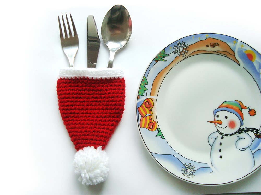 Roter gehäkelter Besteckhalter in Form einer Weihnachtsmütze mit Gabel, Messer und Löffel, daneben ein Teller mit Schneemannmotiv.