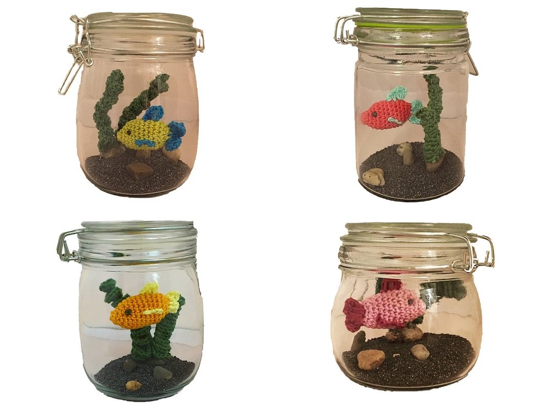 Vier Einmachgläser mit gehäkelten Fischen, Seegras und dunklem Sand