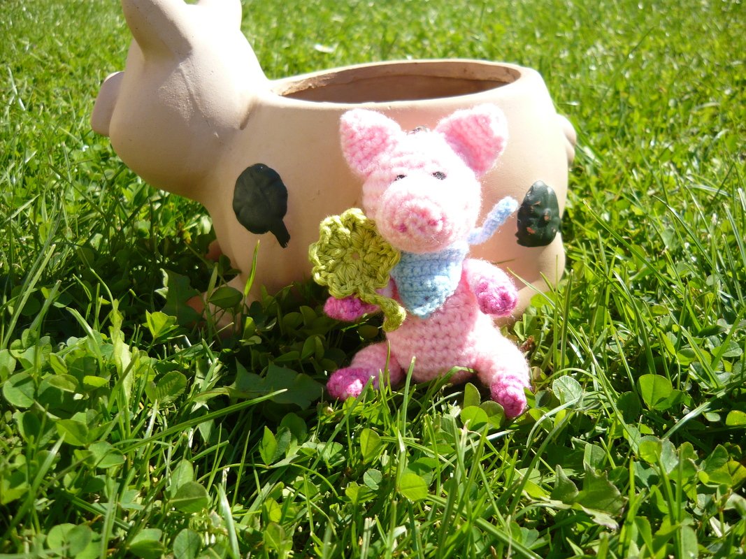 Handgehäkeltes rosa Amigurumi-Schwein mit blauem Halstuch und grünem vierblättrigem Kleeblatt im Gras vor einem beigen Schweine-Pflanztopf.