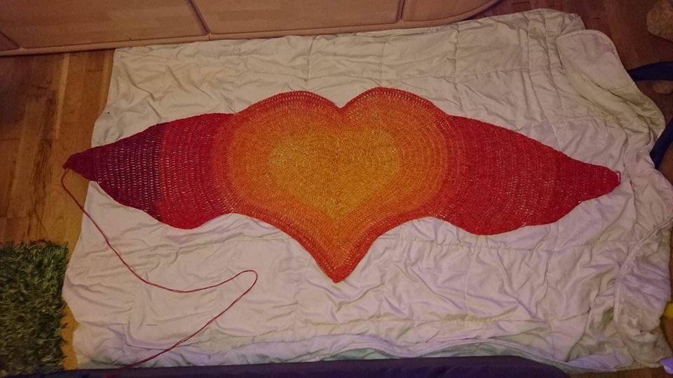 Gehäkelte herzförmige Decke mit Ombre‑Verlauf von Gelb über Orange zu Rot auf heller Unterlage ausgebreitet.