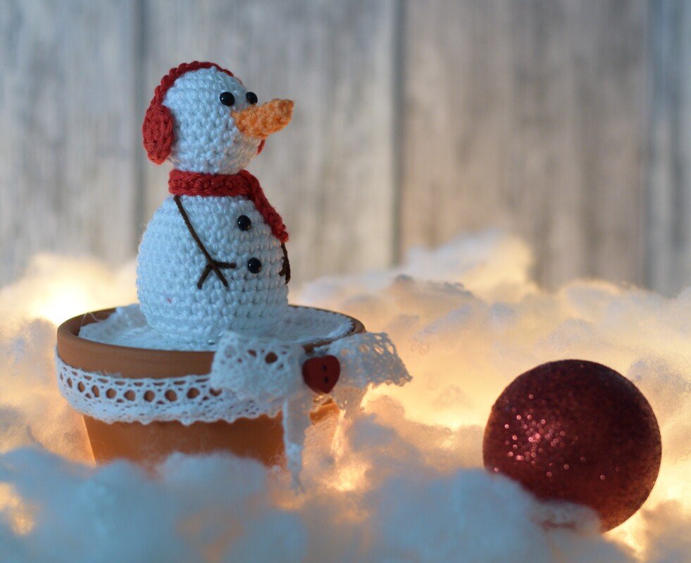 Gehäkelter weißer Schneemann mit rotem Schal und oranger Nase in einem mit Spitze geschmückten Terrakottatopf, umgeben von Lichterglanz und einer roten Christbaumkugel