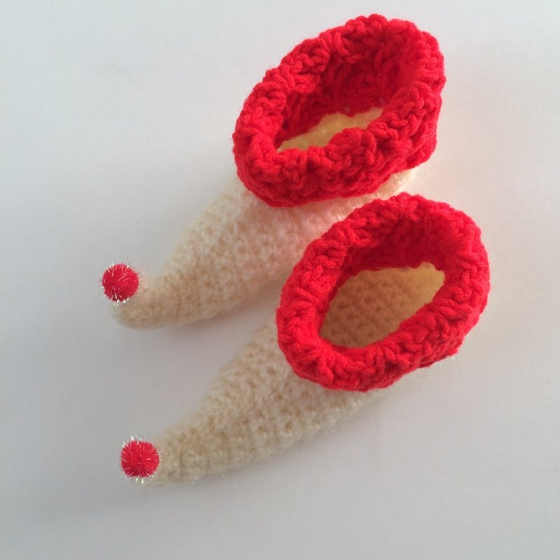 Cream crocheted baby elf shoes with red cuffs and red pom-pom tips on a white background