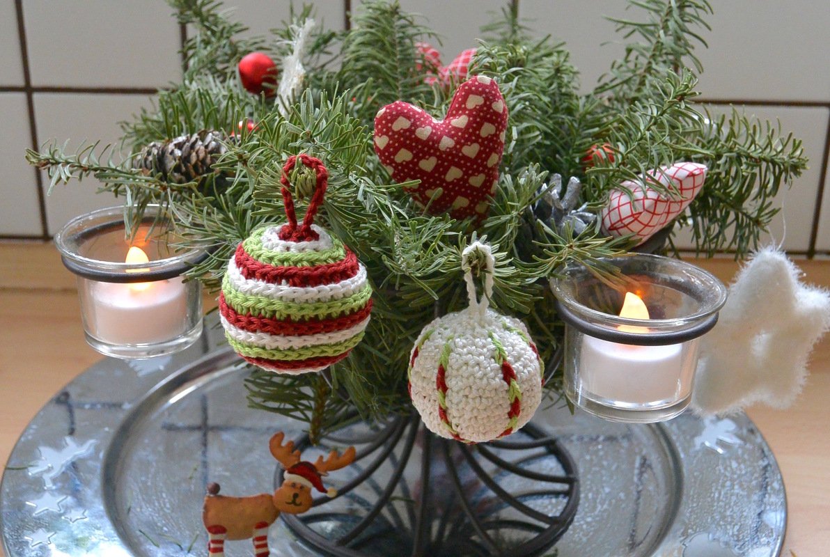 Tischgesteck aus Tannenzweigen mit gehäkelten Weihnachtskugeln, Stoffherz, Kiefernzapfen und zwei Kerzen im Glas