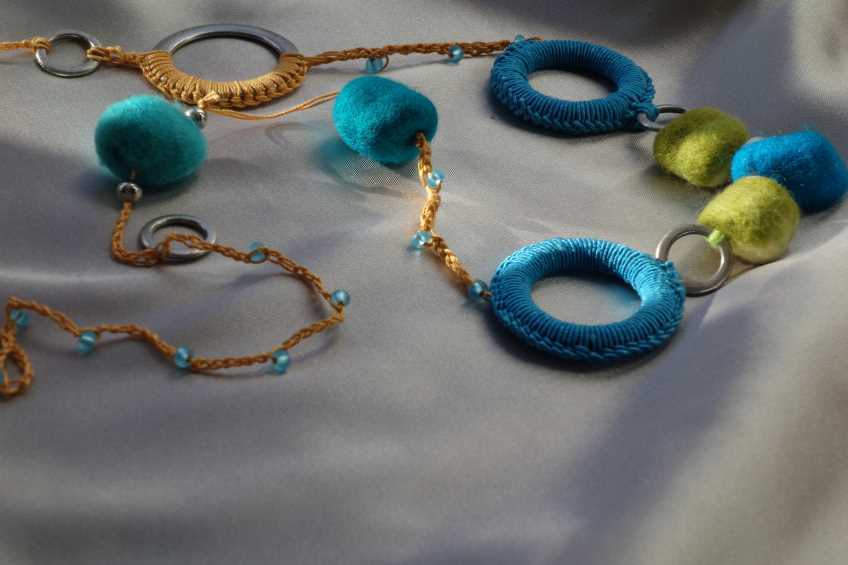 Halskette mit türkisen und limettengrünen Filzbällen, gehäkelten Ringen und geflochtenem Band mit blauen Perlen auf hellem Stoff.