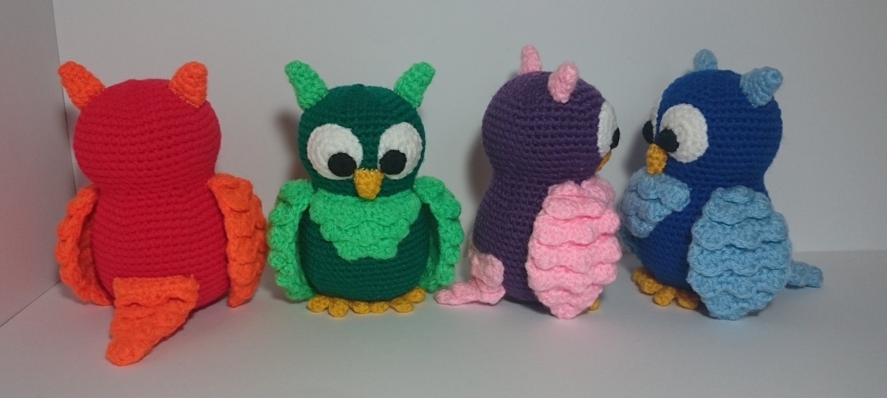 Vier gehäkelte Amigurumi‑Eulen in Rot, Grün, Violett und Blau nebeneinander