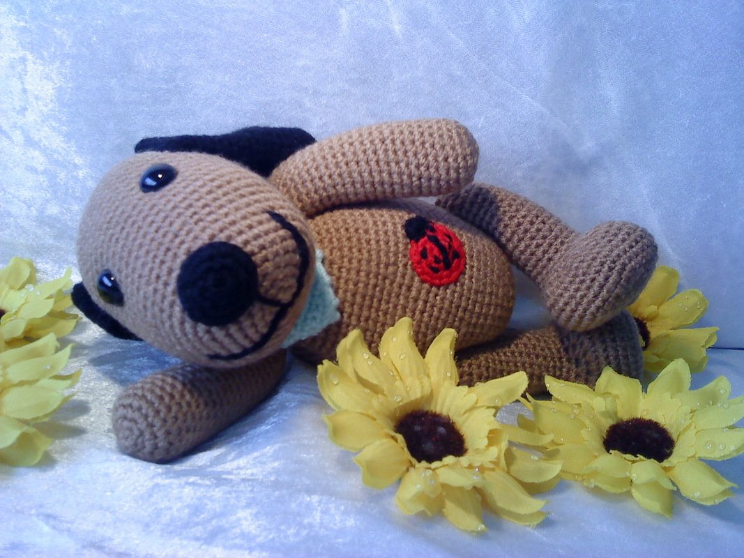 Beiger gehäkelter Amigurumi-Hund mit schwarzer Nase und Ohren, rotem Marienkäfer auf dem Bauch, umgeben von gelben Sonnenblumen.