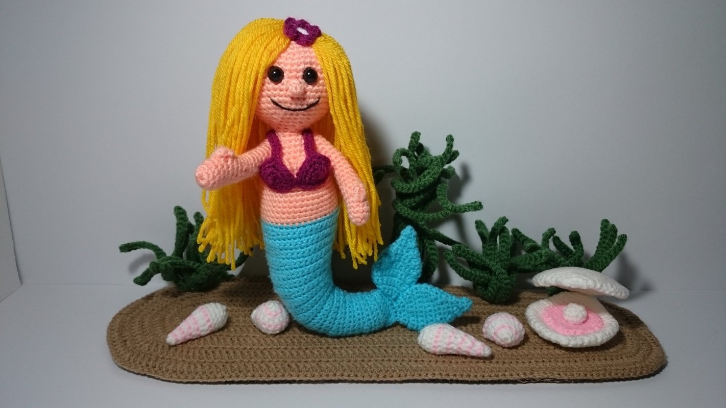 Handgehäkelte Meerjungfrau-Amigurumi mit gelbem Haar, türkisfarbenem Schwanz, Muscheln und grünem Seegras auf einer braunen Häkelunterlage