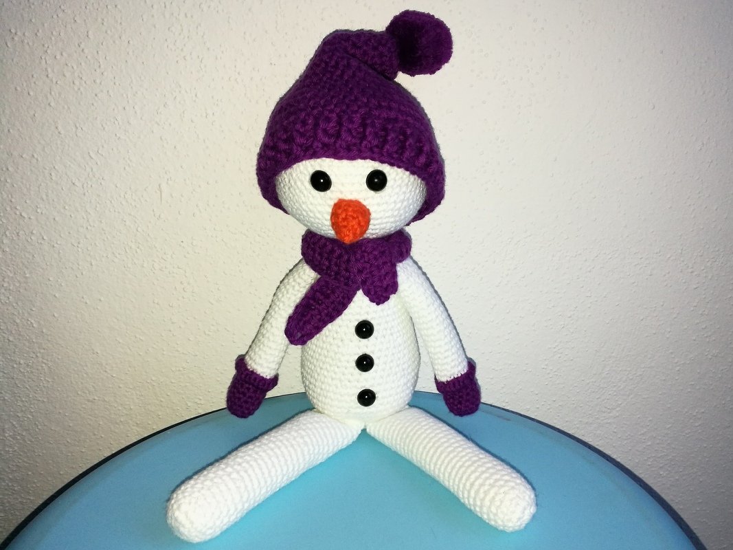 Gehäkelter weißer Schneemann mit lila Mütze und Schal auf blauem Untergrund