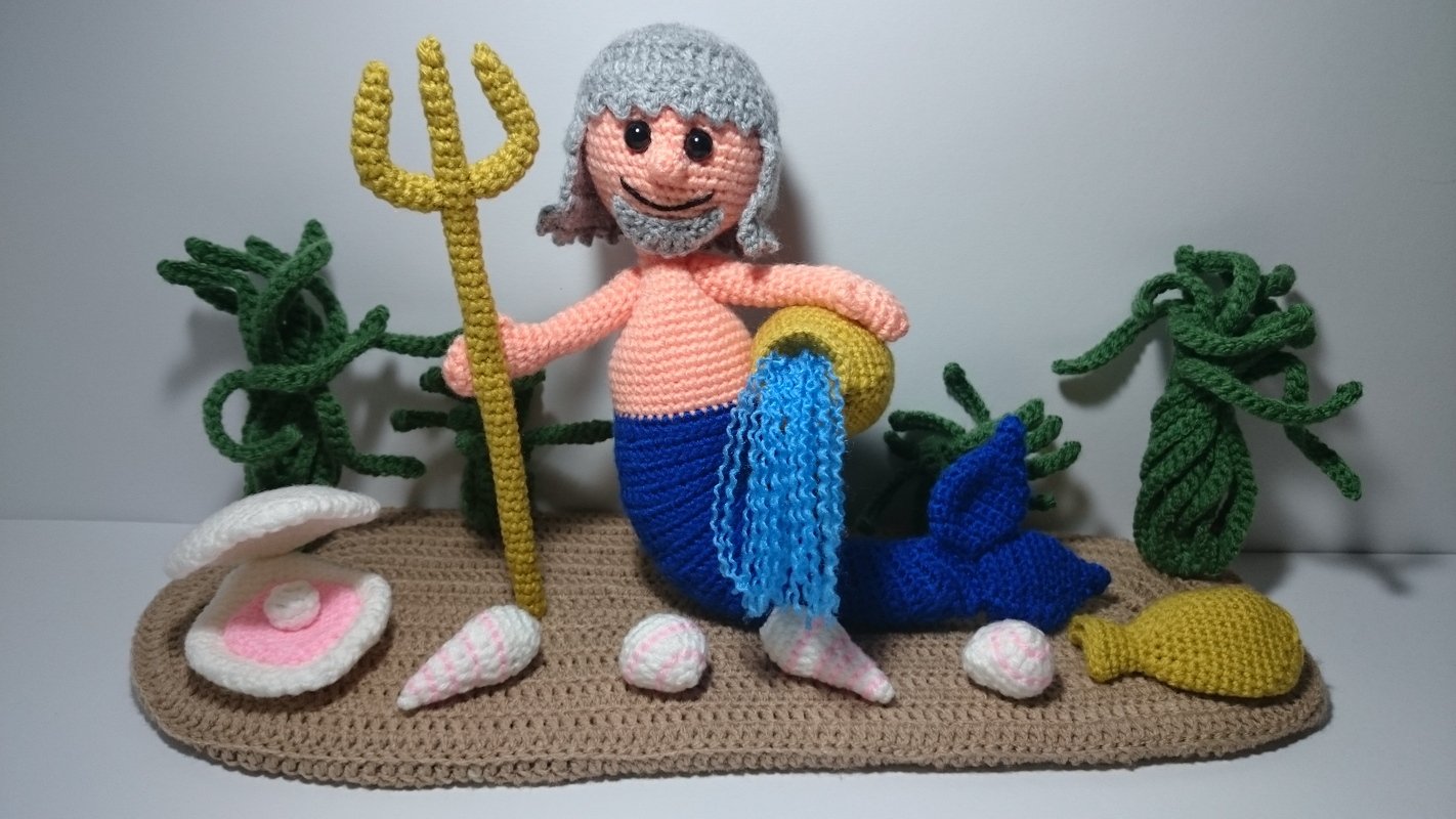 Gehäkelte Meermann-Figur mit grauem Haar, gelbem Dreizack, Muschel mit Perle, Seegras und Fisch auf Häkelunterlage.
