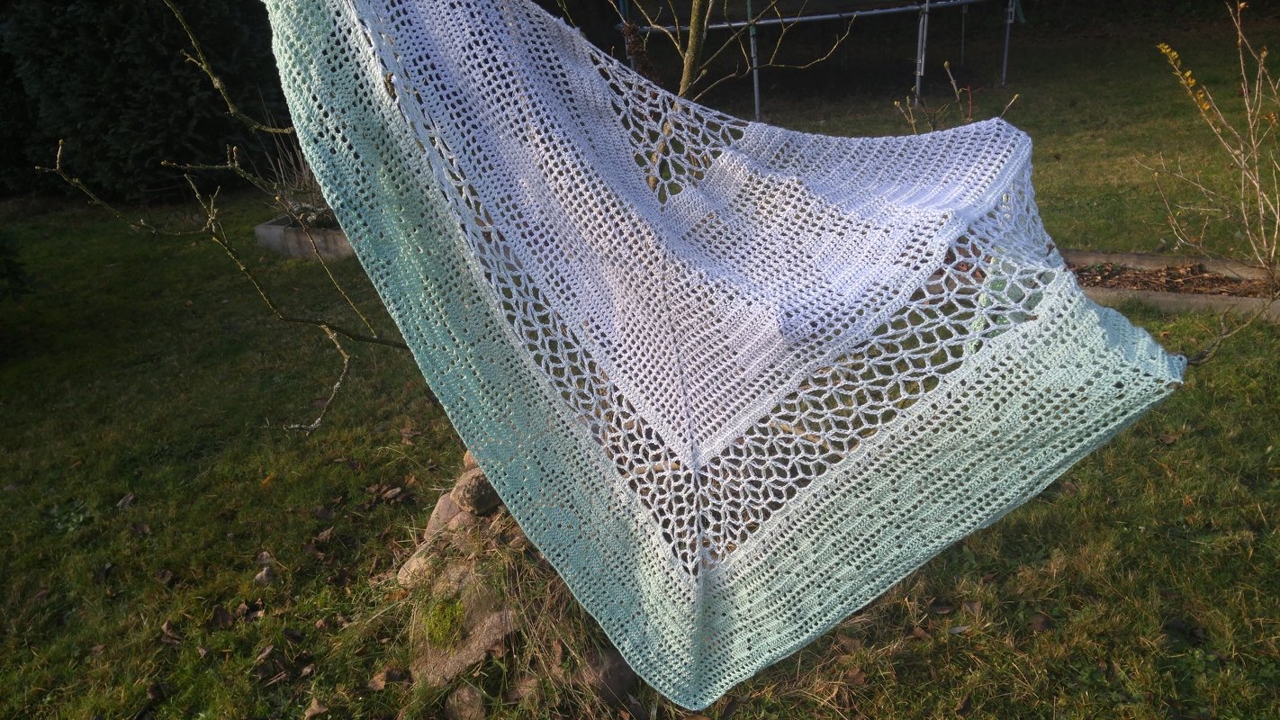 Gehäkeltes Dreieckstuch mit Hellblau-zu-Mintgrün-Verlauf und Spitzen-Gittermuster, an einem Zweig im Freien aufgehängt.