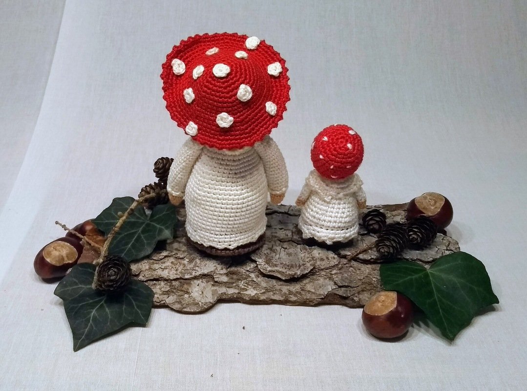 Rückansicht von zwei gehäkelten Fliegenpilz-Puppen auf Baumrinde, dekoriert mit Efeu, Kastanien und Zapfen.