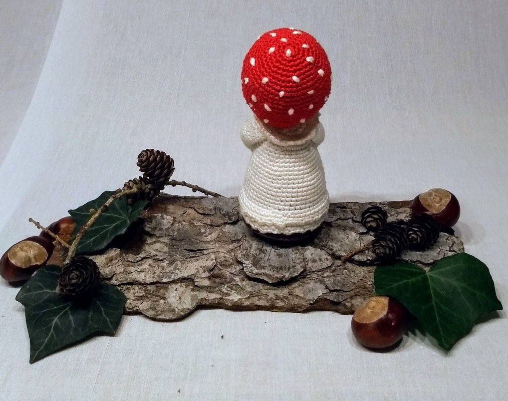 Amigurumi-Häkelpuppe mit roter Pilzmütze und weißem Kleid auf Rindenstück mit Kastanien, Efeu und Zapfen