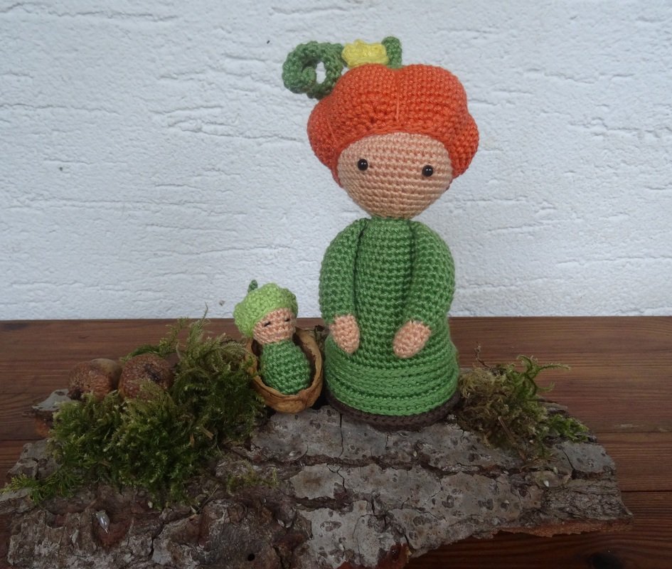 Amigurumi‑Mutter mit orangefarbenem Kopfschmuck und Baby in Nussschale auf Baumrinde mit Moos