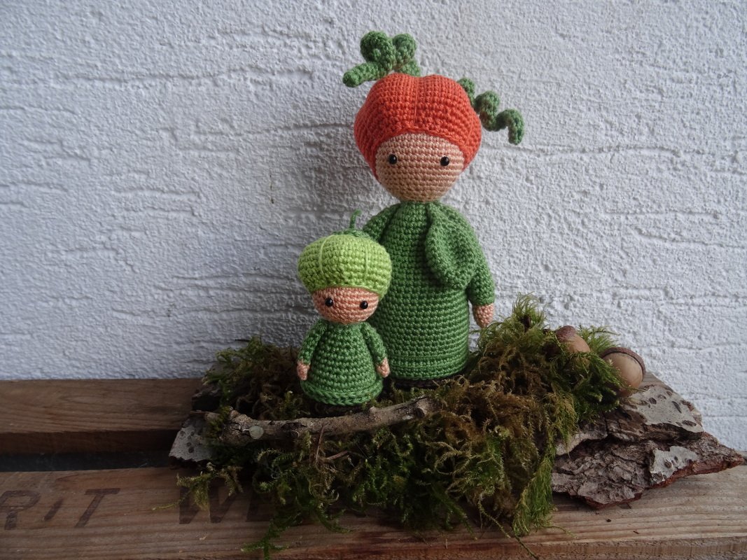 Nahaufnahme zweier gehäkelter Amigurumi-Puppen: größere mit orangefarbener Kürbismütze, kleinere mit hellgrüner Mütze, beide in grünen Kleidern auf Moos und Baumrinde vor weißer Wand