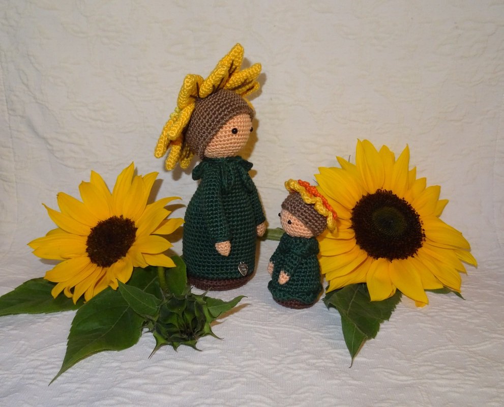 Zwei gehäkelte Amigurumi-Puppen mit Sonnenblumenhüten zwischen Sonnenblumen auf weißem Hintergrund.