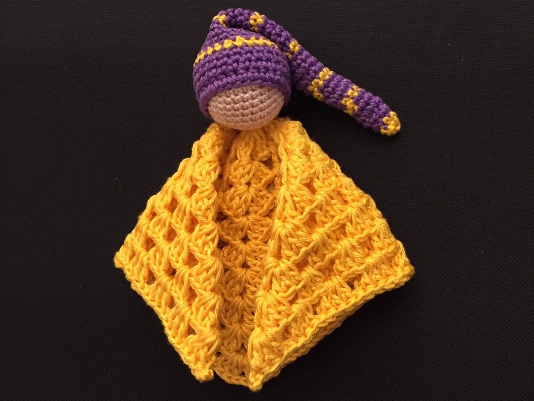 Gehäkeltes gelbes Schmusetuch mit Amigurumi-Kopf und lila-gelber Zipfelmütze auf dunklem Hintergrund