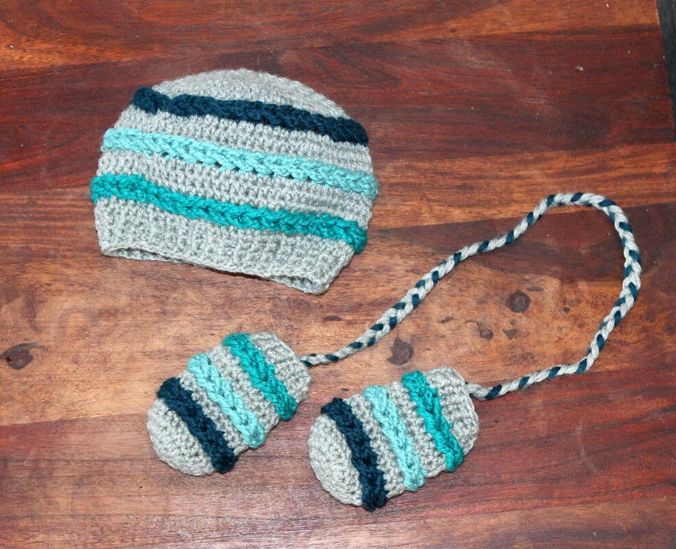 Gehäkelte Babymütze und Fäustlinge in Grau mit türkisen und dunkelblauen Streifen auf Holztisch