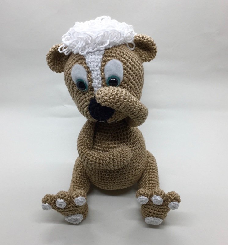 Gehäkelter hellbrauner Amigurumi-Bär mit weißer Lockenpartie und weißen Pfotenakzenten, sitzt mit einer Pfote vor dem Mund.