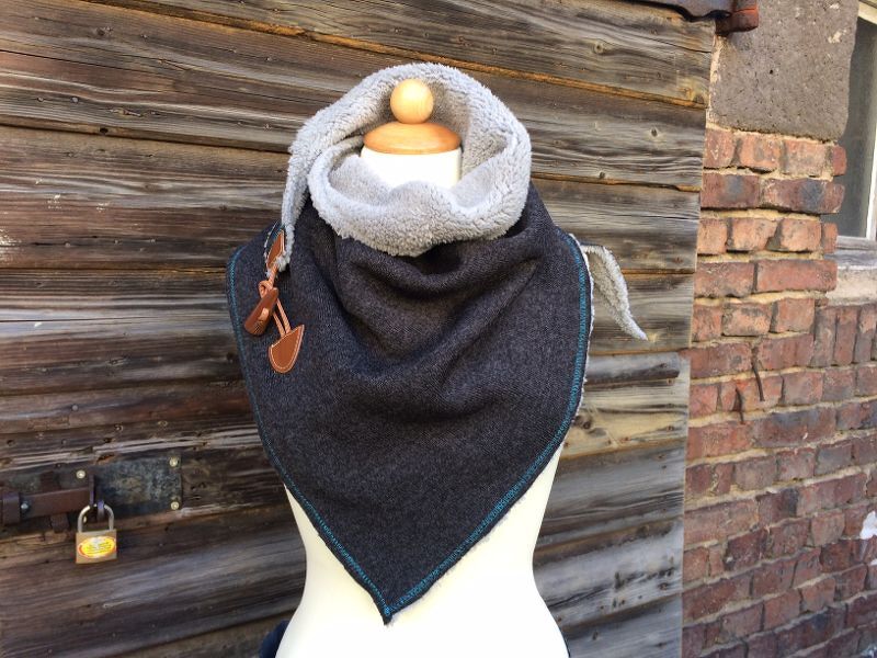 Dunkelgraues Dreieckstuch mit hellgrauem Fellfutter, türkiser Kettelnaht und lederähnlichen Verschlüssen auf Schaufensterpuppe vor Holzfassade