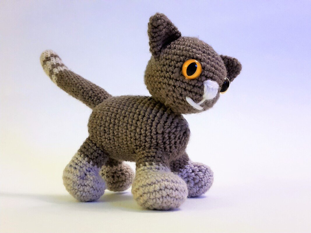 Gehäkelte Amigurumi-Katze braun-grau mit orangefarbenen Augen und gestreiftem Schwanz auf hellem Hintergrund