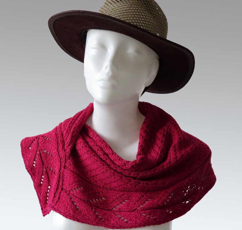 Deep red knitted cowl with lace eyelets and cable pattern draped on a white mannequin wearing a brown wide-brim hat