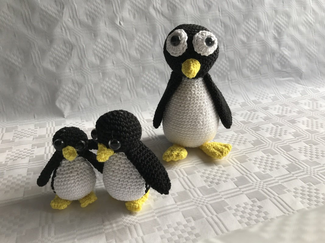 Drei gehäkelte Amigurumi‑Pinguine (zwei kleine, ein großer) auf weißem, gemustertem Stoff