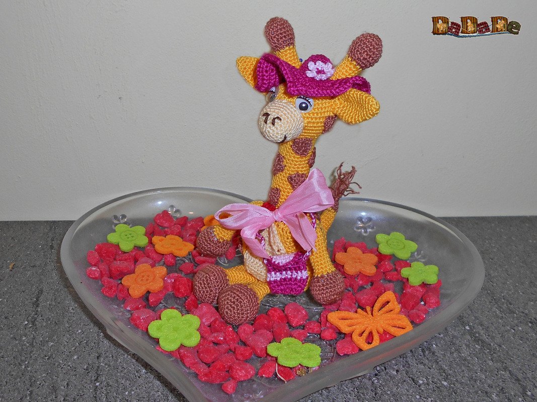 Gehäkelte gelbe Giraffe mit braunen Flecken, rosa Hut und Schleife in einer herzförmigen Schale mit roten Steinen und bunten Blüten und Schmetterlingen.