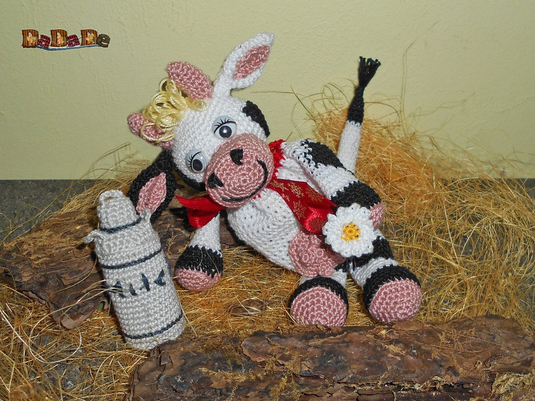 Gehäkelte schwarz-weiße Amigurumi-Kuh mit roter Schleife und Blume neben gehäkelter Flasche auf Stroh
