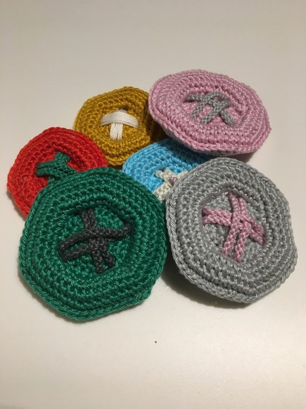 Sechs runde gehäkelte Untersetzer in Rot, Grün, Gelb, Hellblau, Rosa und Grau mit mittigen Kreuzmustern.