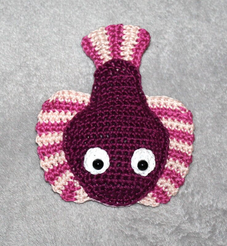 Flaches gehäkeltes Amigurumi-Fischmotiv in Dunkelviolett mit pink-rosa gestreiften Flossen und weißen Augen auf grauem Hintergrund