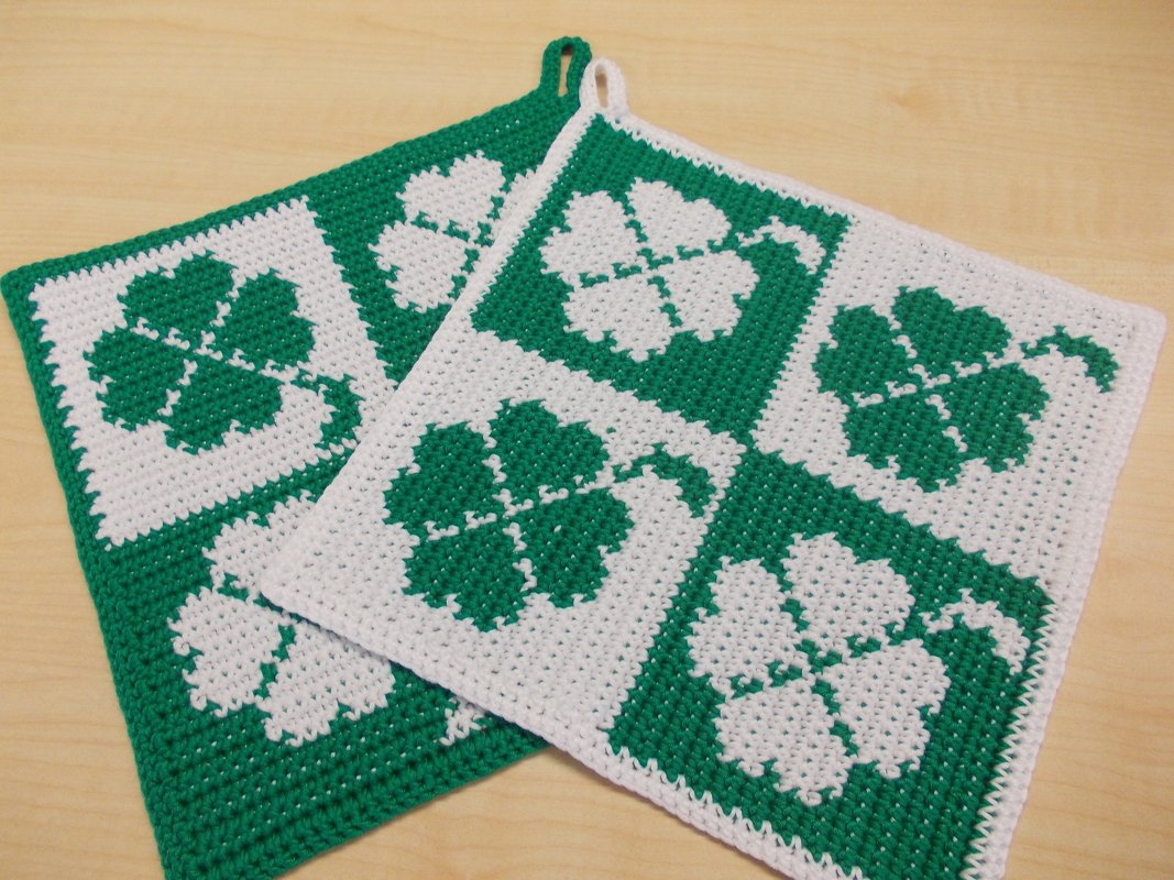 Zwei grün-weiße gehäkelte Topflappen mit Kleeblattmotiven auf Holzuntergrund.
