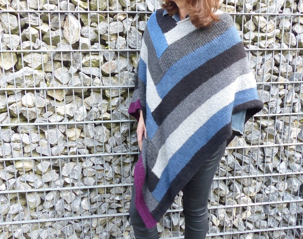 Gestrickter Poncho mit diagonalen Streifen in Blau, Grau, Schwarz und Weiß vor einer Gabionenwand