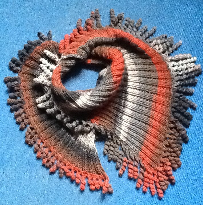 Gestrickter Loop Schal mit Streifen in Grau, Braun, Orange und Weiß und Noppenrand auf blauem Untergrund