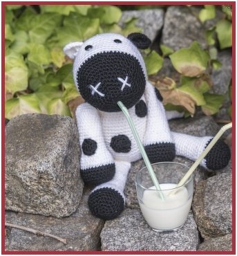 Weiß‑schwarze handgehäkelte Amigurumi‑Kuh sitzend auf Steinen neben einem Glas Milch mit Strohhalm