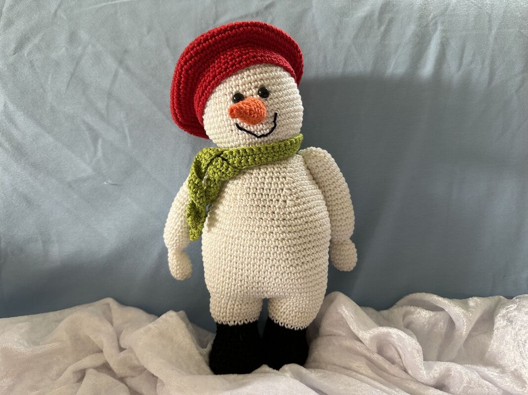 Gehäkelter Amigurumi-Schneemann als Häkelpuppe mit roter Mütze, grünem Schal, orangefarbener Nase und schwarzen Füßen