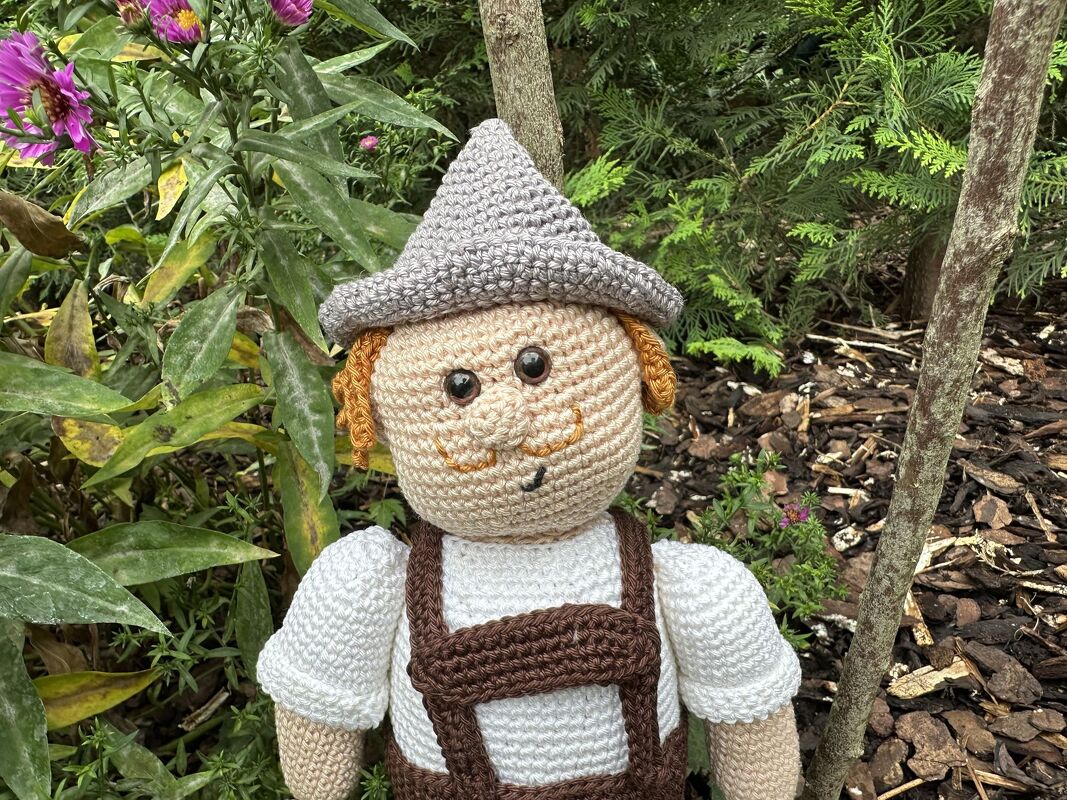 Gehäkelte Amigurumi-Puppe mit brauner Hose, Hosenträgern und grauem Spitzhut im Garten.