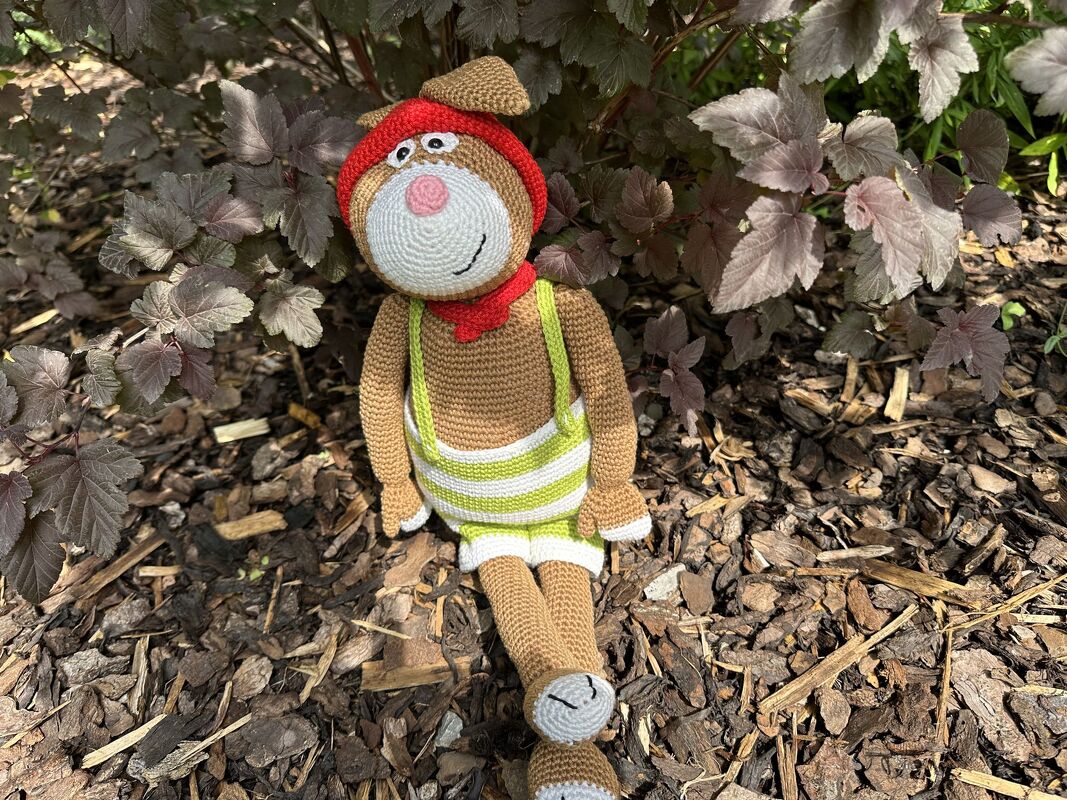 Braune Amigurumi-Tierfigur mit roter Kappe, Halstuch und grün-weiß gestreifter Latzhose, sitzend auf Rindenmulch