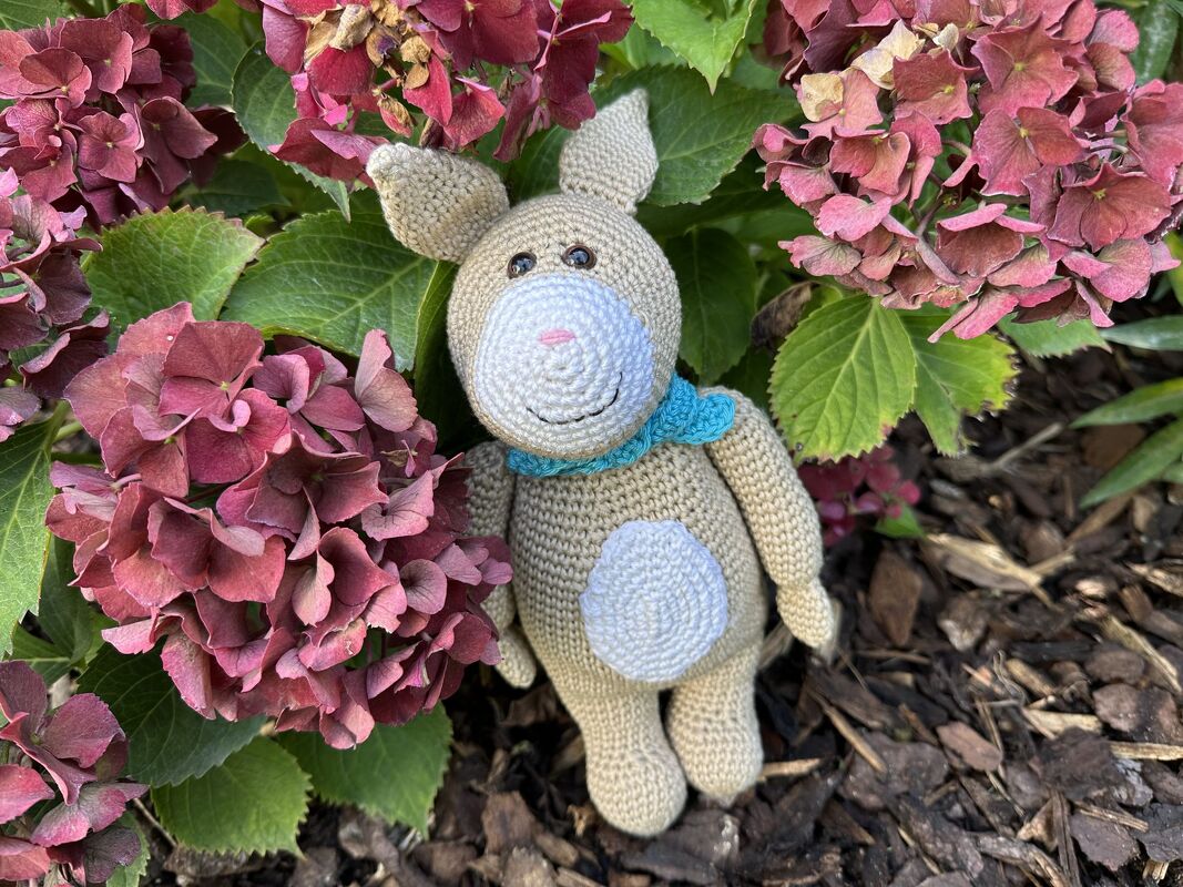 Beiger gehäkelter Amigurumi-Hase mit hellblauem Schal zwischen rosa-roten Hortensienblüten.