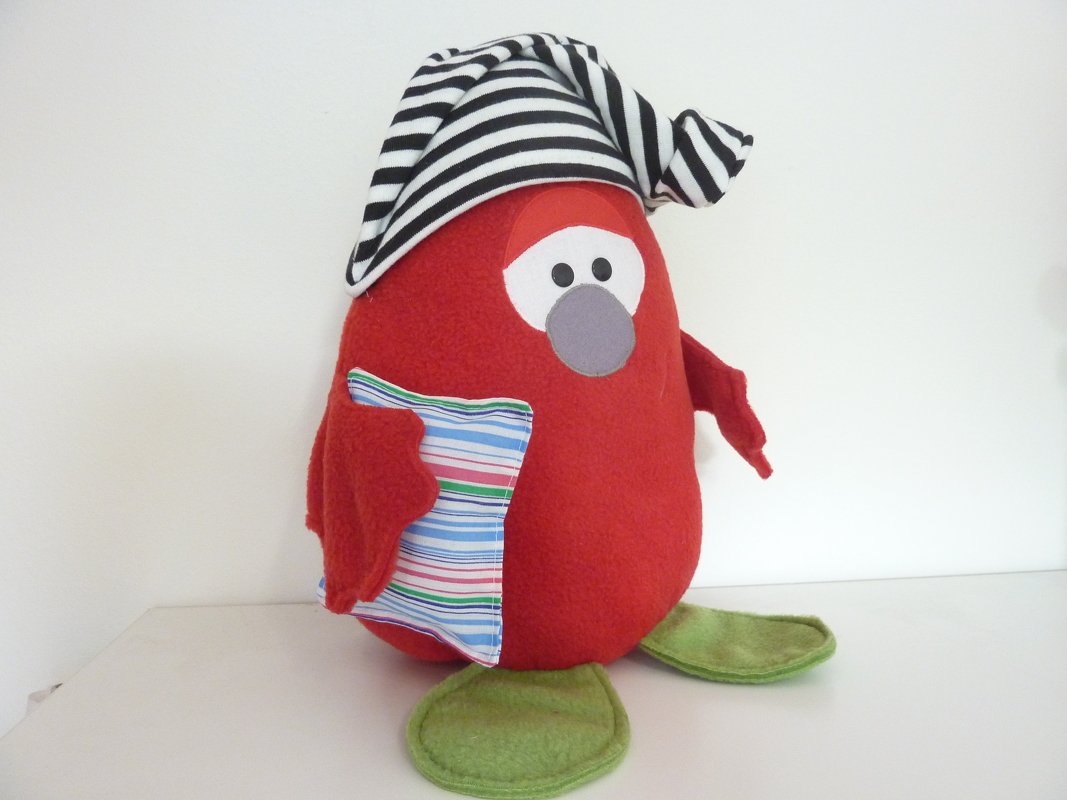 Rotes rundes Kuscheltier mit schwarz-weiß gestreifter Mütze, angenähten Flügeln, gestreiftem Mini-Kissen und grünen Füßen
