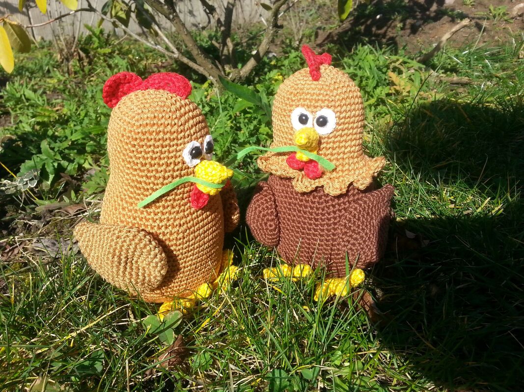 Zwei Amigurumi-Hühner mit roten Kämmen und gelben Schnäbeln sitzen im Gras.