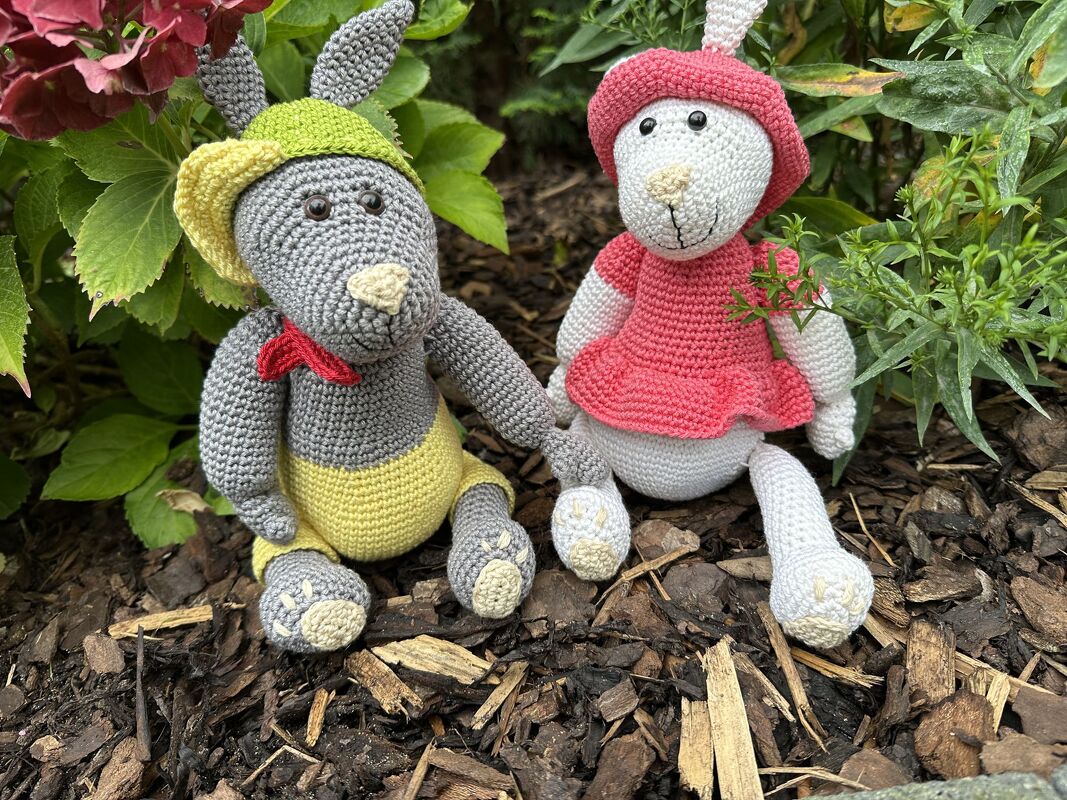 Amigurumi-Hasenpaar mit Outfits: grauer Hase mit gelber Hose, rotem Halstuch und grün-gelbem Hut, weißer Hase mit rosa Kleid und Hut.