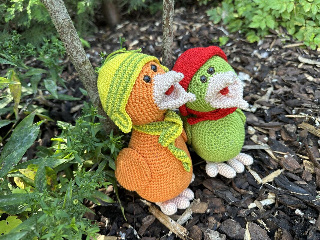 Zwei gehäkelte Amigurumi-Vögel, orange mit gelb-grüner Mütze und Schal sowie grün mit roter Mütze und Schal, auf Rindenmulch