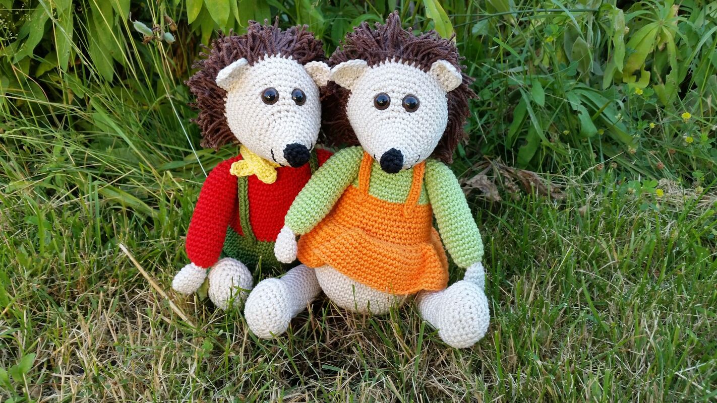 Zwei gehäkelte Igel-Amigurumi mit braunen Stacheln und bunten Kleidern sitzen im Gras