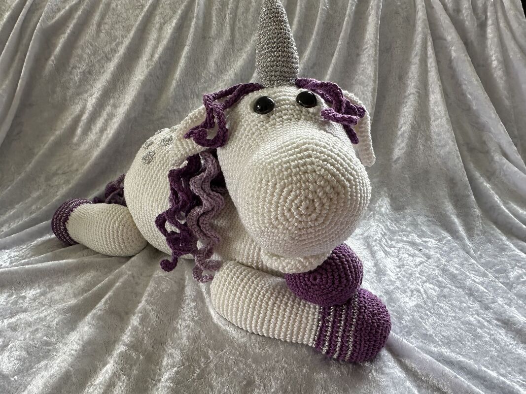 Weißes gehäkeltes Amigurumi-Einhorn mit violetter Mähne und silbernem Horn auf hellem Stoff