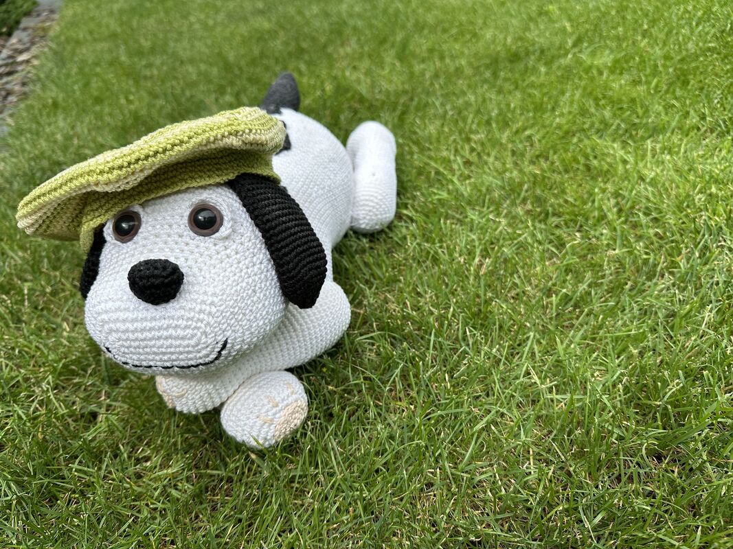 Weißer Amigurumi-Hund mit schwarzen Ohren und grüner Schirmmütze auf Gras