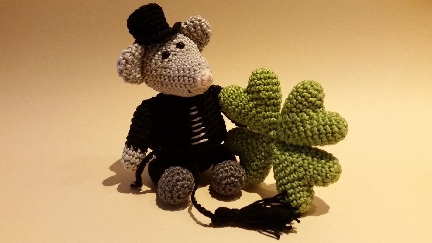 Gehäkelte Amigurumi‑Maus mit Zylinder neben einem grünen vierblättrigen Kleeblatt.