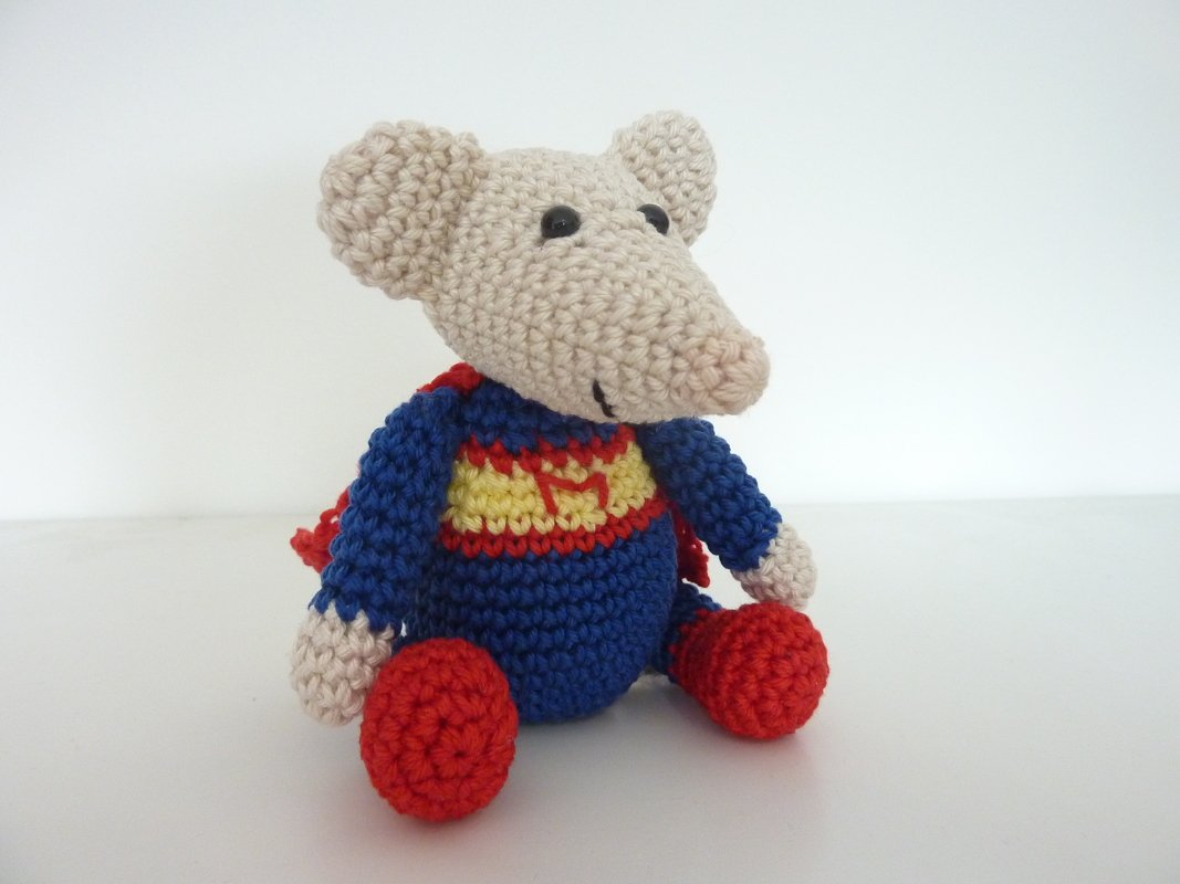 Beige gehäkelte Amigurumi‑Maus mit blauem Oberteil, rotem Umhang und roten Füßen, sitzend vor weißem Hintergrund