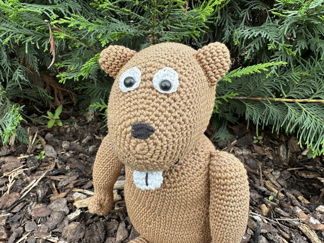 Brauner gehäkelter Amigurumi-Biber mit großen Augen und weißen Nagezähnen vor grünen Zweigen