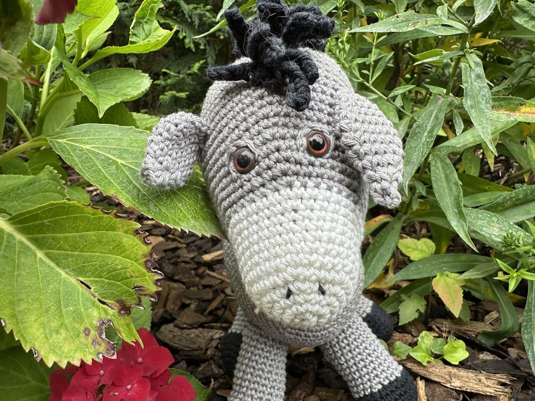Grau-weißer gehäkelter Amigurumi-Esel mit schwarzer Mähne zwischen grünen Gartenblättern