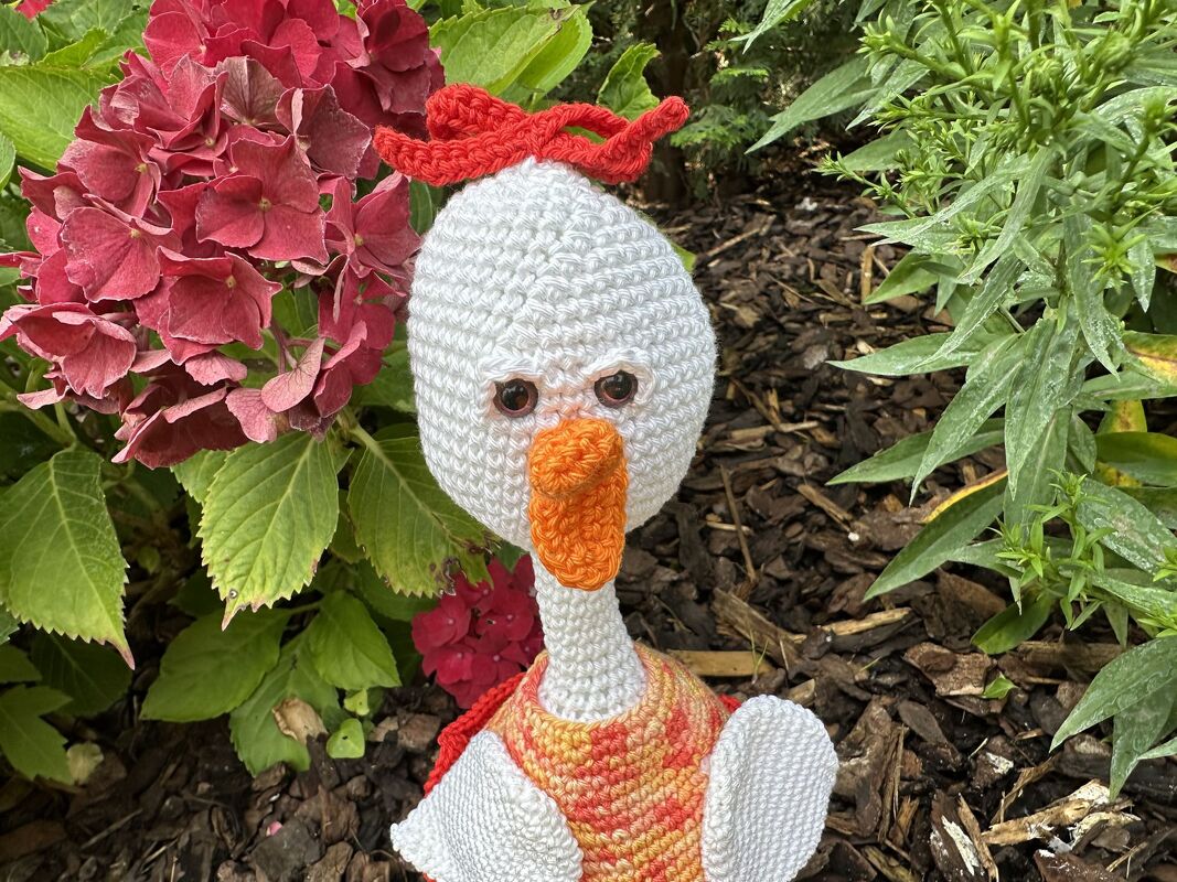 Weiß gehäkelte Amigurumi-Gans mit orange Schnabel und roter Schleife vor Gartenpflanzen
